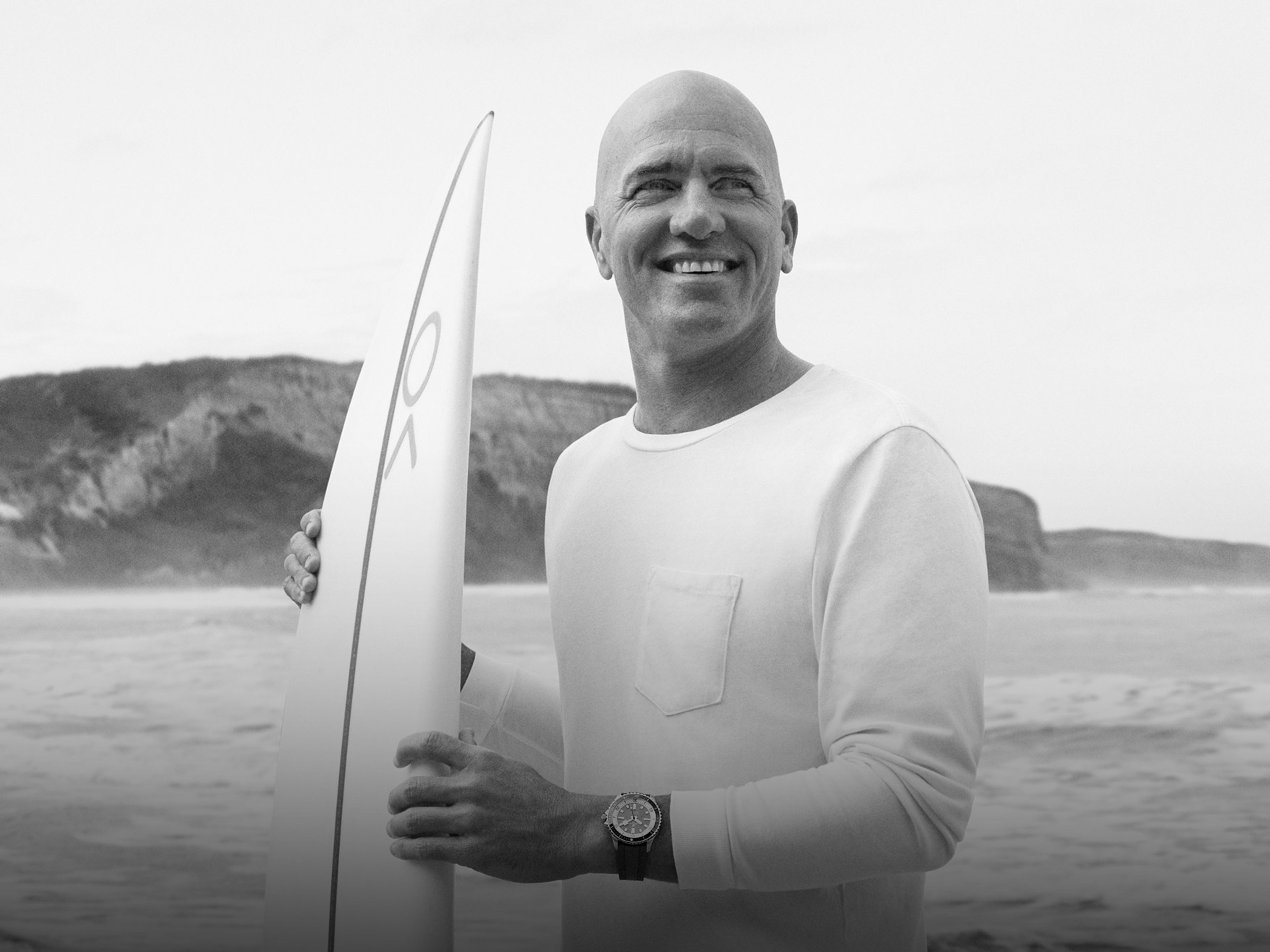 {generated} A surfer holding a board on a rocky shoreline, wearing a Breitling watch that highlights its sporty coastal style