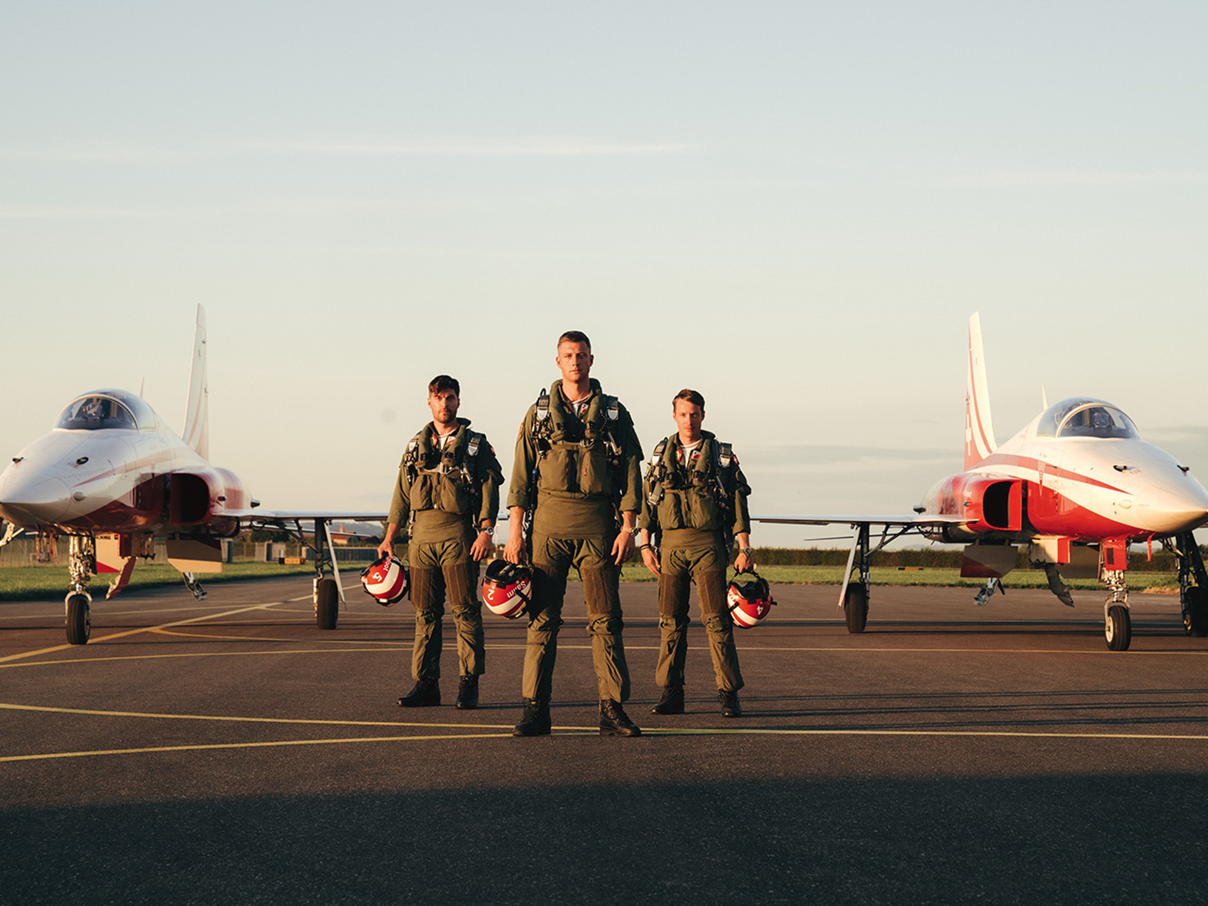 {generated} Three pilots in flight suits stand between two red‑white jets at sunset, expressing Breitling’s aviation heritage.