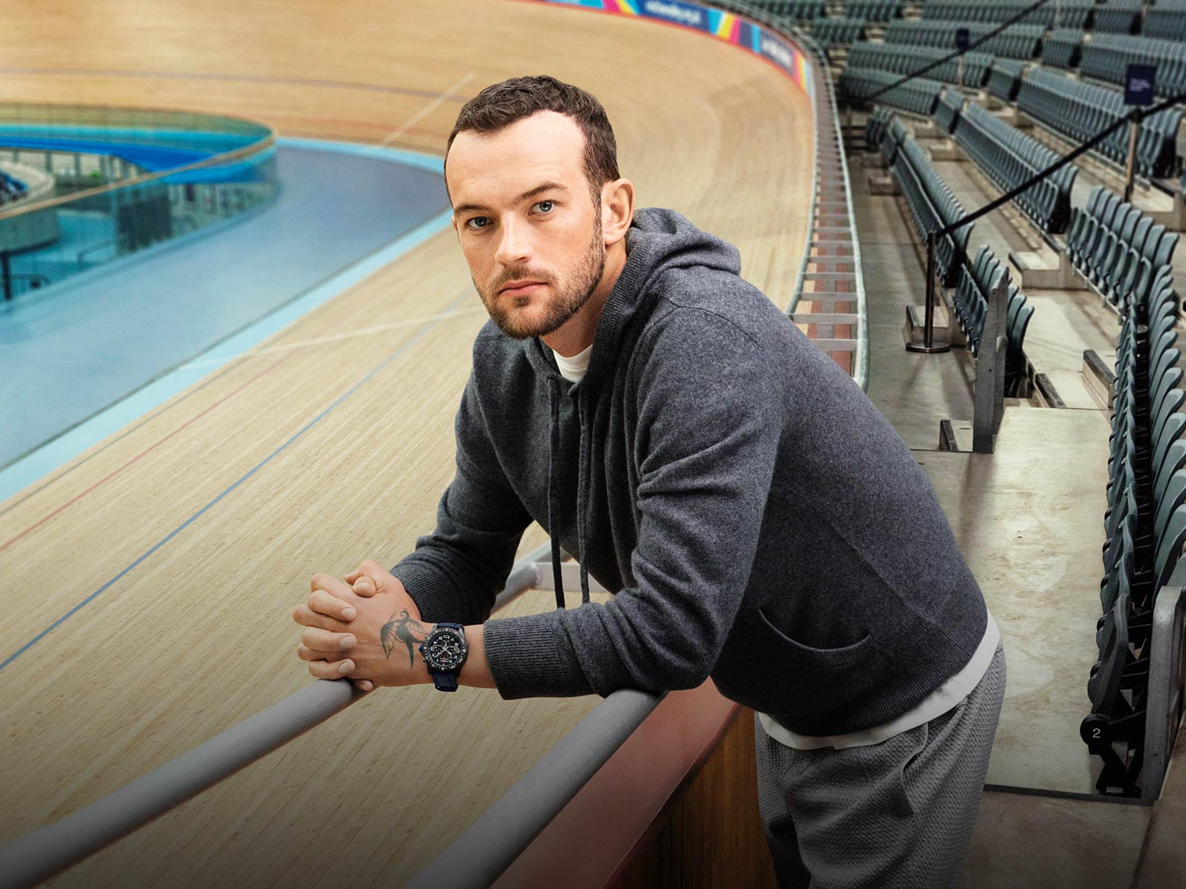 {generated} A man at a velodrome rail wearing a dark hoodie and a bold Breitling watch, shown in a calm athletic setting