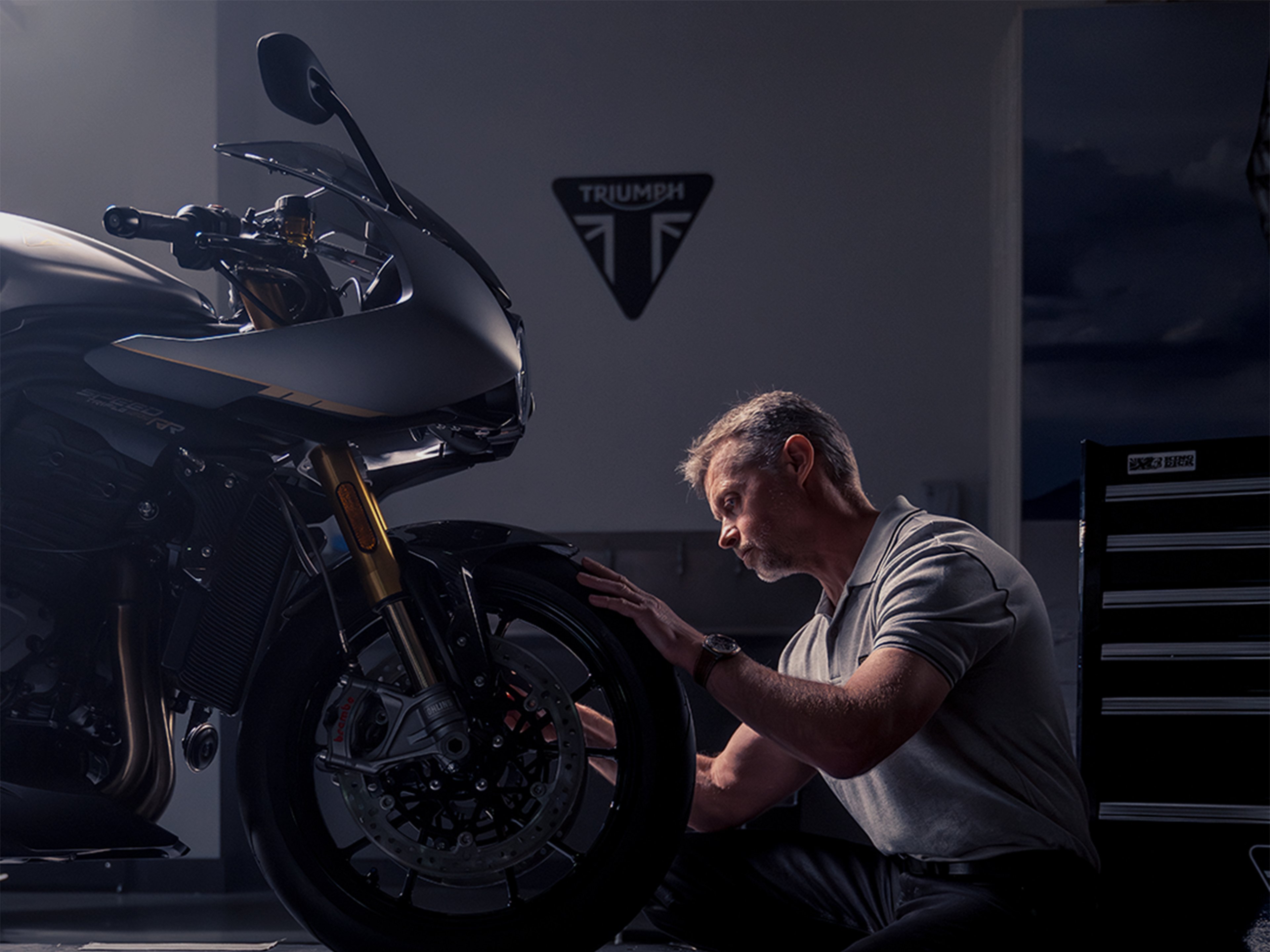 Man with Breitling watch in focus kneels in front of Triumph motorbike