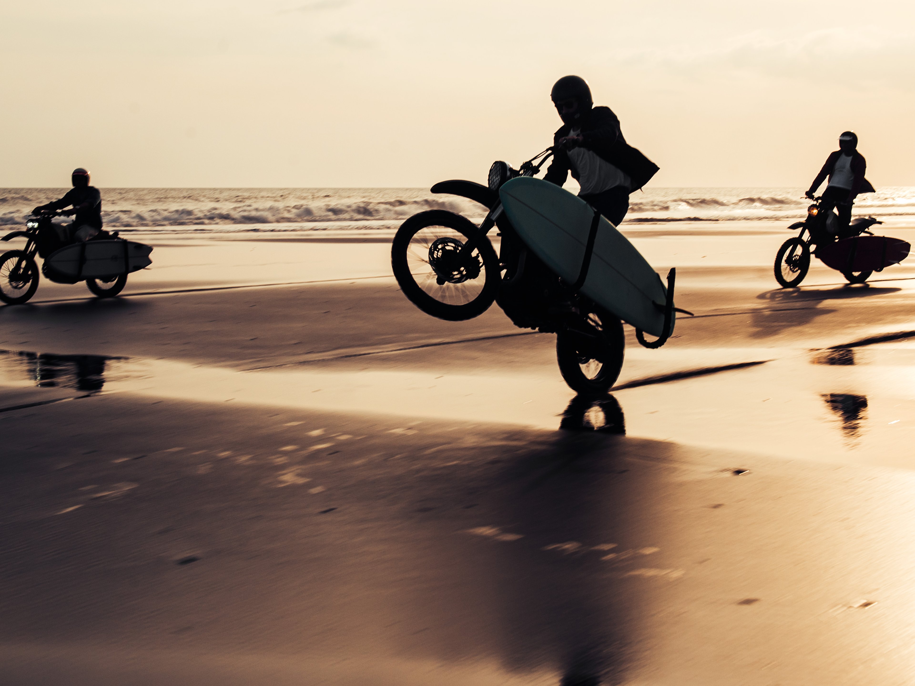 Three Deus Ex Machina motorbikes with surfboards in tow ride through the surf on the beach at sunset 