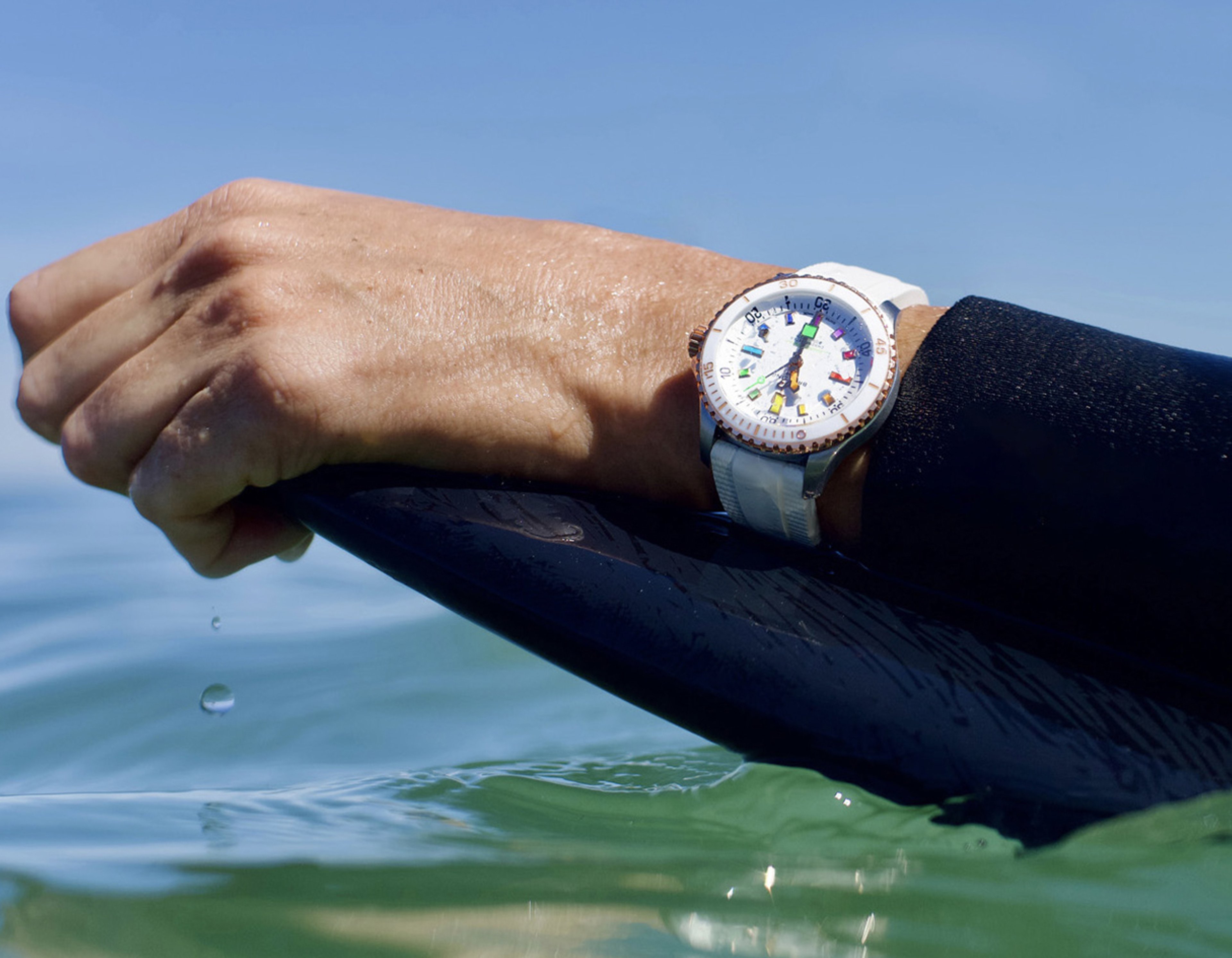 {generated} Un polso con Breitling Superocean dal cinturino bianco afferra una tavola bagnata sopra acqua limpida sotto un cielo blu brillante.