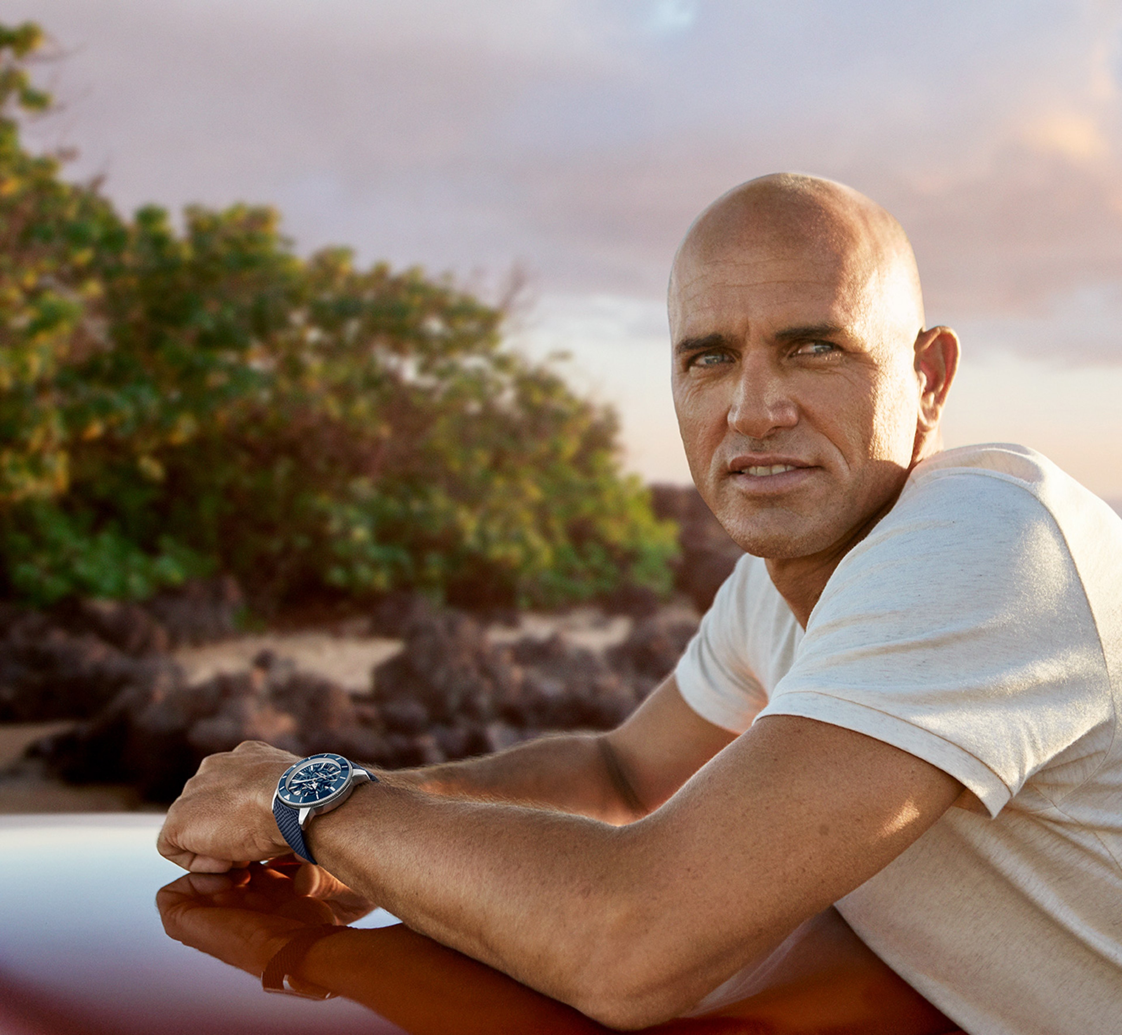 {generated} A man by a coastal landscape rests his arm forward, showcasing a Breitling SOH watch with blue dial and strap in warm light.