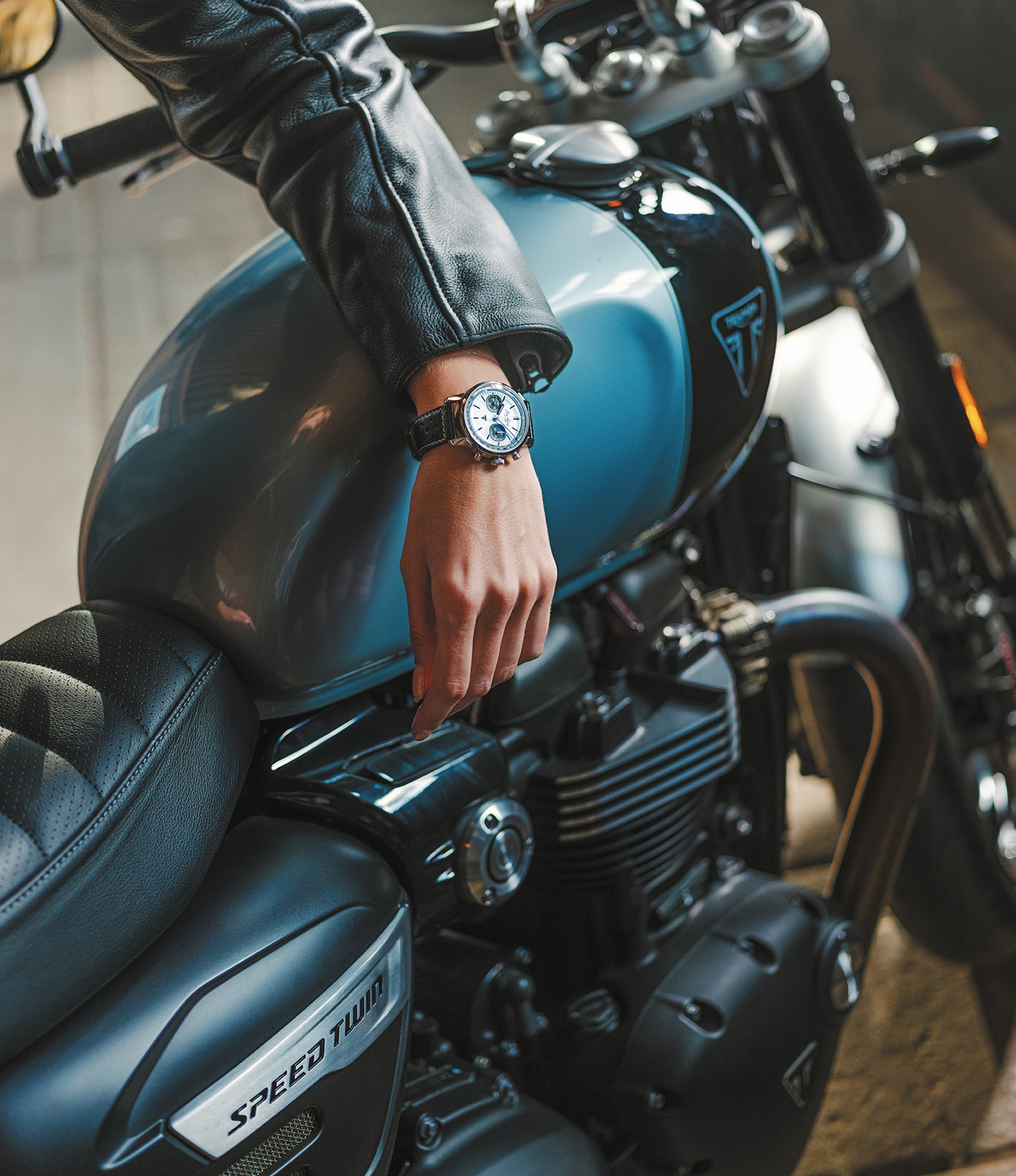 {generated} A rider rests a hand wearing a Breitling Top Time Triumph on a blue Triumph motorcycle, highlighting its bold detailing.