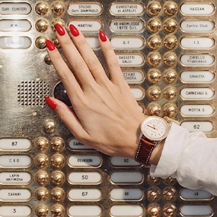 {generated} Eine Hand mit roten Nägeln trägt eine Breitling Uhr und liegt auf einem Messingklingelpanel, betont elegante Details.